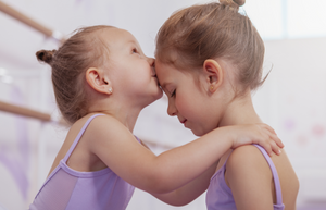 Two young female ballet students wearing lavender leotards are facing each other. One ballet student is kissing the other on the forehead. 