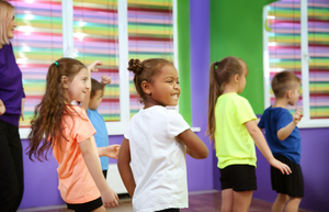 A young dance class filled with males and females. The dancers are wearing bright colored shirts and black shorts. The walls are bright purple with colorful blinds over the windows. 