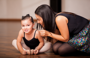 A young female ballet student wearing a black leotard, pink ballet tights and a bow, is sitting on the floor stretching in a "butterfly" position, smiling, and her female dance instructor wearing a black leotard is kneeling beside her, also smiling. 